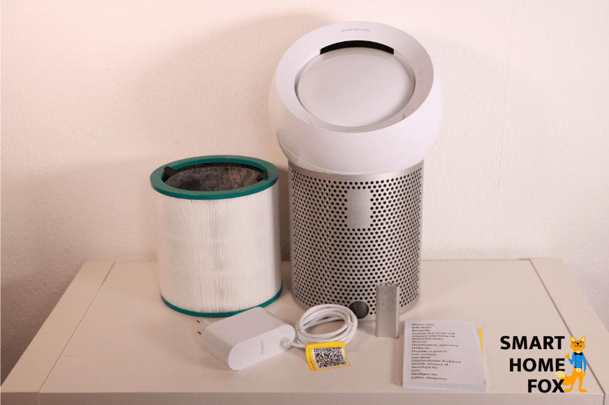 Der Dyson Pure Cool Me BP01 Luftreiniger und Tischventilator steht auf dem Regal. Daneben befindet sich die Glas-HEPA- und Aktivkohlefilter-Einheit, die Fernbedienung, das Stromkabel sowie die Bedienungsanleitung.